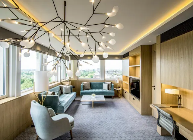 A hotel suite living area with two teal sofas, a branch-style pendant light, a wood-panelled wall with a television and a desk, and windows looking out over a city with trees.