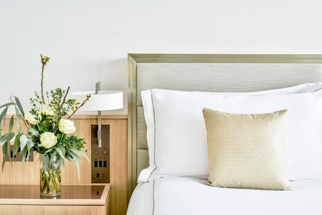 A made bed with white linen and a gold patterned cushion sits beside a bedside table holding a vase of white roses and a wall-mounted lamp.