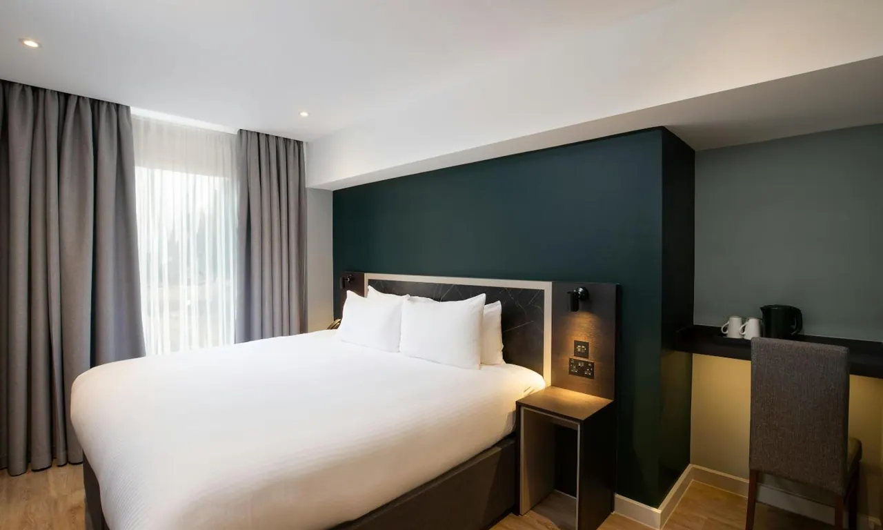 A hotel bedroom with a double bed under white bedding, a dark green accent wall, grey curtains and a desk with a kettle and two cups.