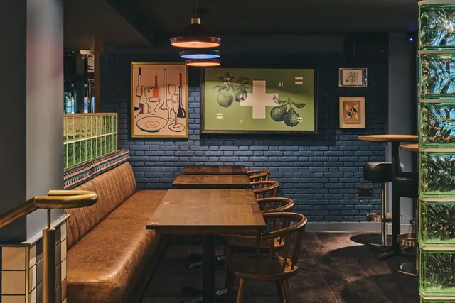 A dimly lit dining area with a tan leather banquette, wooden tables and chairs, framed artwork on a dark blue brick wall, and a green glass block partition on the right.