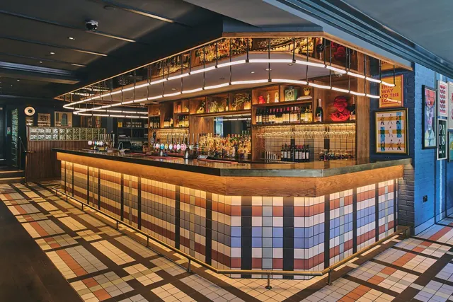 A bar counter with a mosaic-tiled front, beer taps, backlit bottle shelves and hanging glassware, with framed posters on the wall beside it.