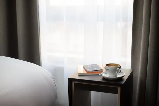 A coffee cup on a saucer and a book sit on a dark bedside table beside a bed with white bedding, sheer curtains and a window behind.
