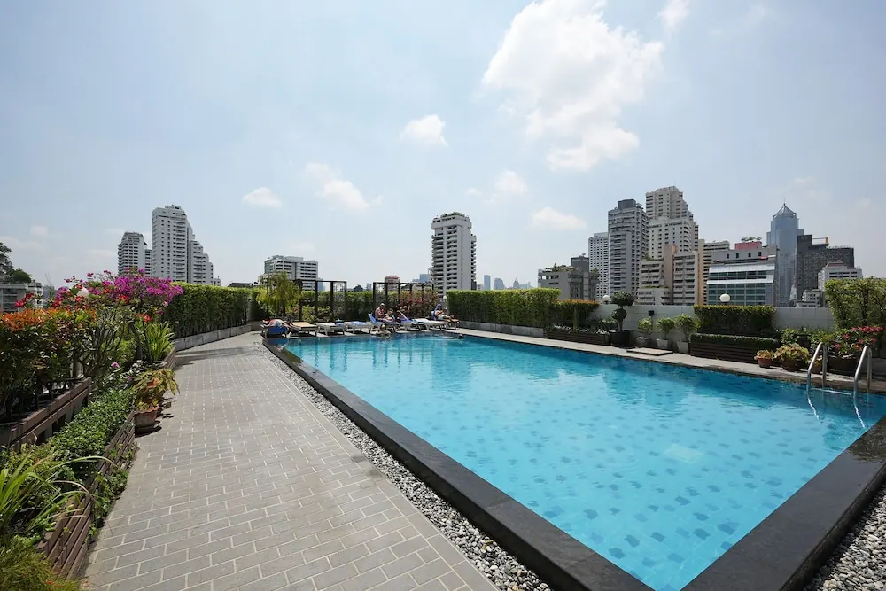 A rooftop swimming pool with sun loungers, planters and high-rise buildings in the background under a partly cloudy sky.