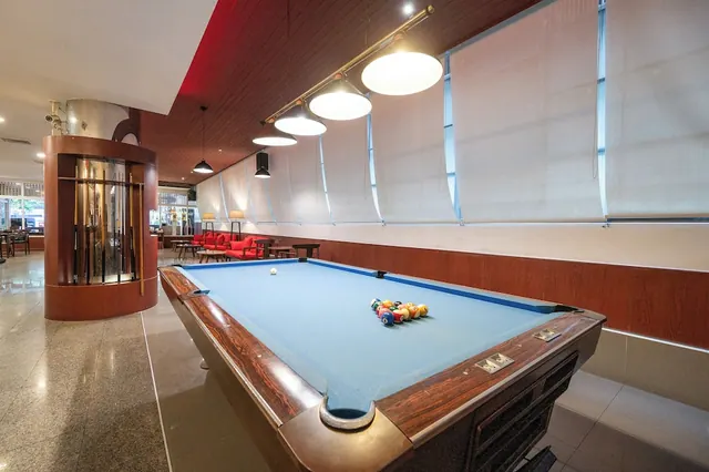 A pool table with blue felt and coloured balls sits in a lounge area with red armchairs in the background and a cue rack beside it.