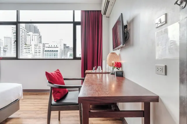 A hotel room with a wooden desk, a red-cushioned armchair beside a window looking onto city buildings, a wall-mounted television and an air conditioner above the door.