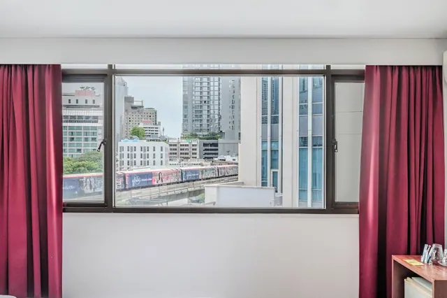 A window with maroon curtains looks out over high-rise buildings and an elevated train line.