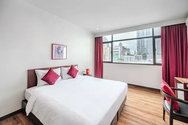 A bedroom with a made king bed with red cushions, a window onto city high-rises, and a desk with a chair on the right.
