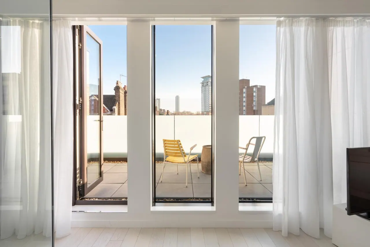 Tall glass doors open onto a paved balcony with a yellow chair, a white chair and a wicker stool, with residential towers and rooftops visible beyond the railing under a clear sky.