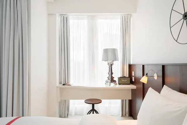 A bedroom with a made bed, a white desk and wooden stool in front of a window with sheer curtains, and a table lamp on the desk.