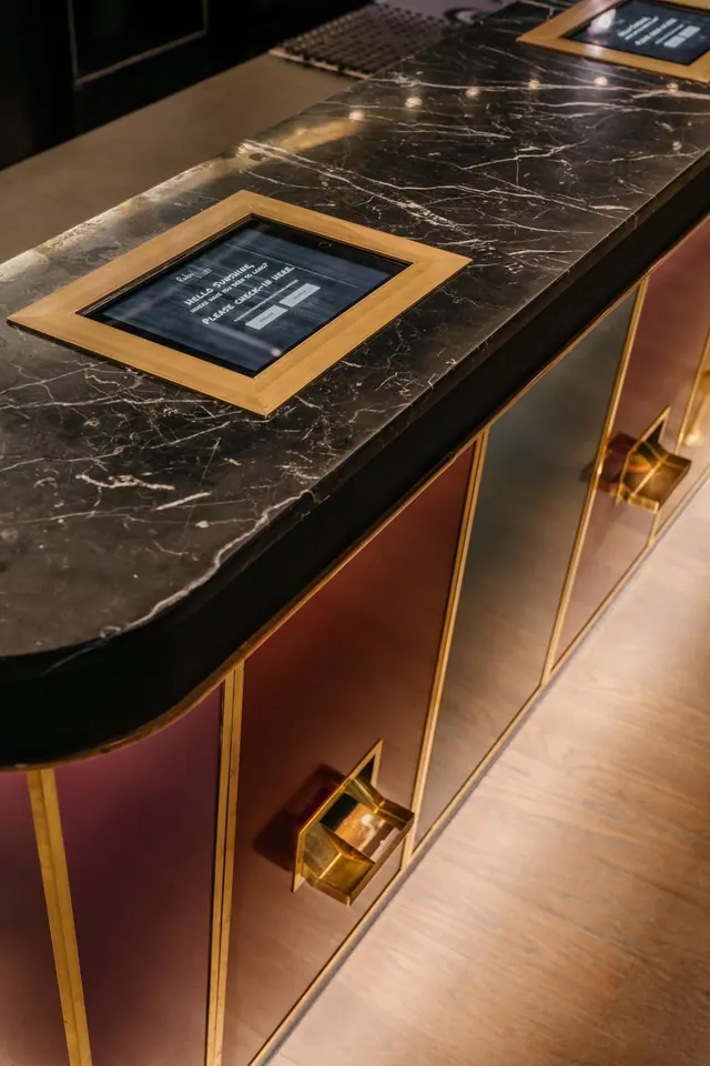 A black marble reception counter with two embedded screens showing check-in messages, brass trim and mauve panels, and a wooden floor.