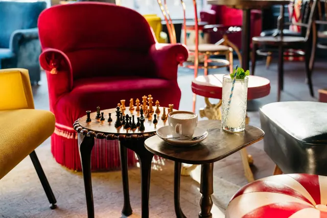 A red velvet armchair, a chess set on a small wooden table, and a coffee cup and cocktail glass on a side table in a lounge.