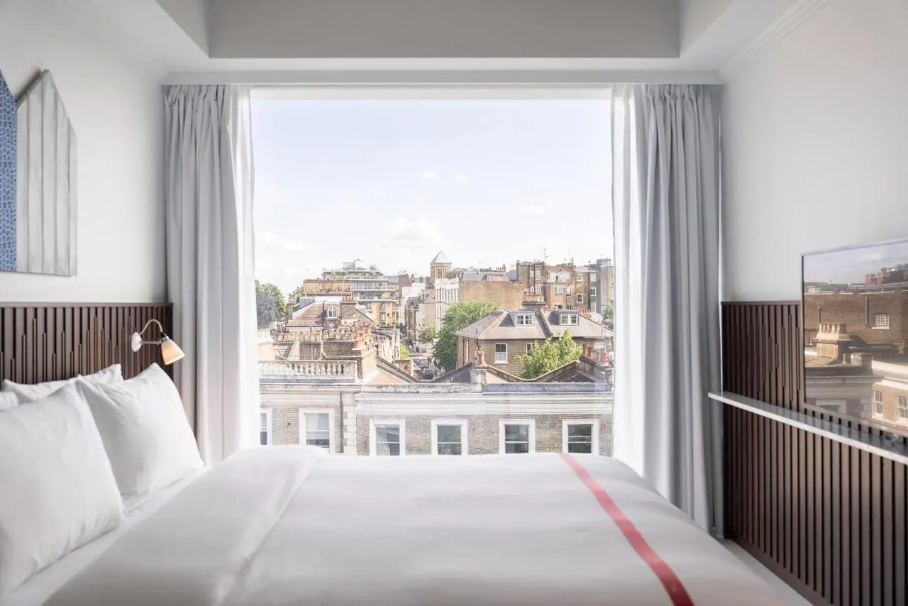 A double bed with white linen and a red stripe faces a window looking out over rooftops and buildings in a city.