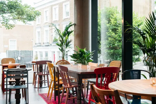 A dining room with mismatched painted wooden chairs and wooden tables, with large windows looking onto a street lined with trees and brick terraced houses.