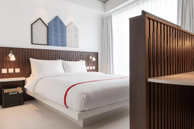 A bedroom with a made double bed with white linen and a red stripe, a wood-slatted headboard wall, sheer curtains over a large window, and a Marshall amplifier on the bedside table.