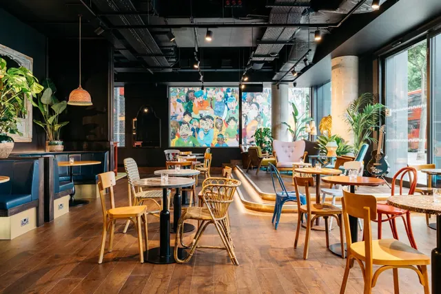 A dining room with wooden tables and chairs, a large colourful mural on the back wall, plants, and floor-to-ceiling windows looking out onto the street.