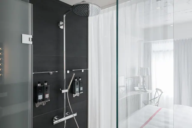 A walk-in shower with a rain head and handheld spray sits beside a glass partition that looks through into a bedroom with a table lamp and a window.
