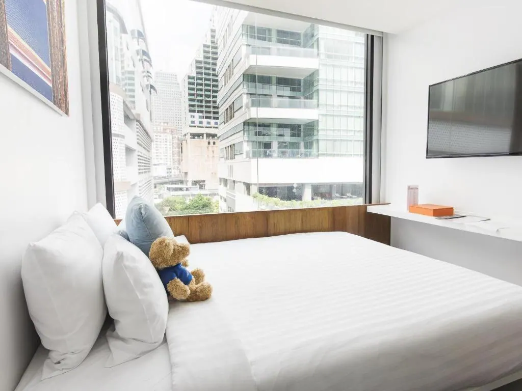 A made bed with white linen and a teddy bear sits beside a large window showing city high-rise buildings, with a wall-mounted television and a white ledge desk on the right.