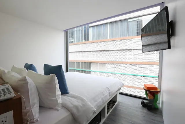 A made-up bed with white linen and blue cushions sits beside a floor-to-ceiling window that looks onto a perforated facade of a neighbouring building, with a wall-mounted television on the right.
