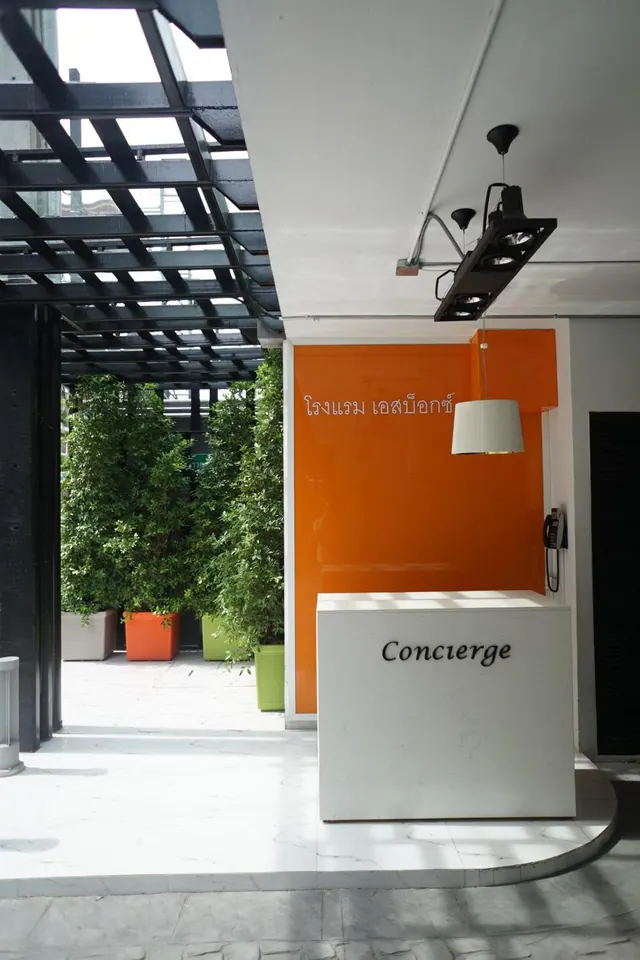 A white concierge desk stands in front of an orange wall with Thai script, with potted plants and a metal pergola overhead.