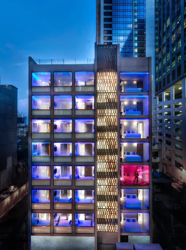 Exterior of a multi-storey hotel building at dusk, with illuminated guest rooms with beds visible through a grid of windows and a taller tower behind.