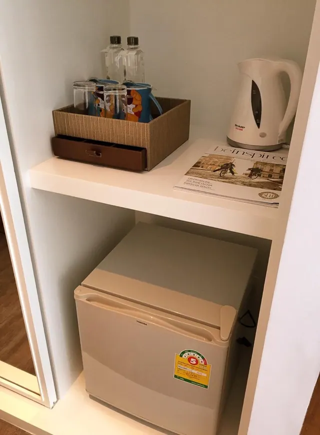 A built-in cabinet nook holds an electric kettle, glasses in a woven basket, bottled water and a magazine, with a small fridge on the lower shelf.