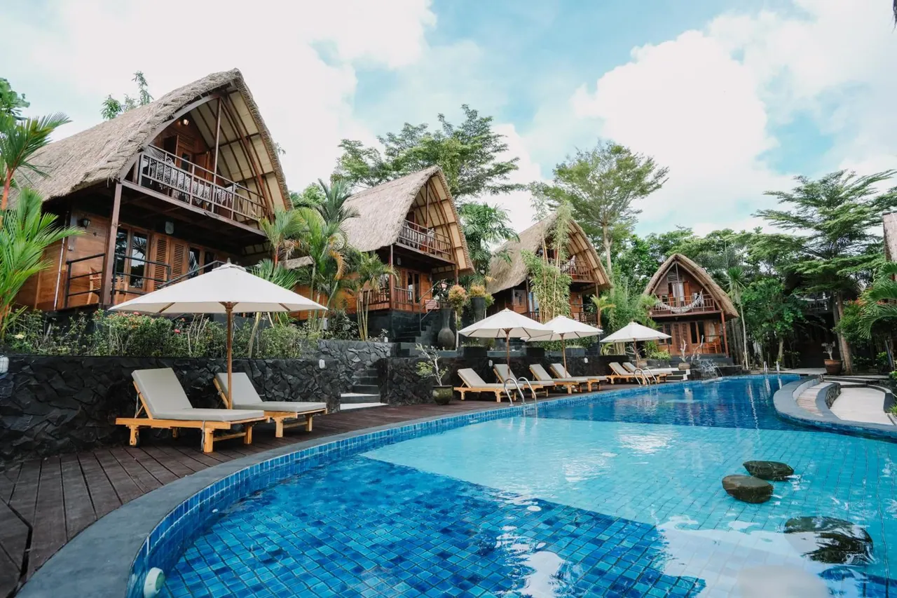 A curved swimming pool with sun loungers under white parasols, with thatched-roof bungalows and tall trees behind.