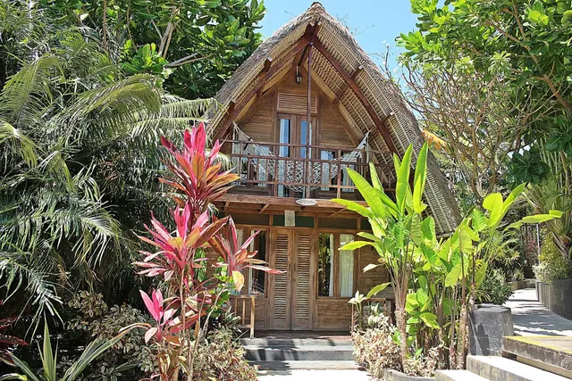 A two-storey thatched-roof wooden bungalow with a balcony and hanging seat, set among tropical plants with stone steps leading to the entrance.