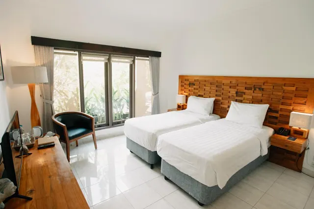 A bedroom with two single beds with white linen and a wooden headboard, a wooden desk with a television and kettle, and large windows looking out onto tropical plants.