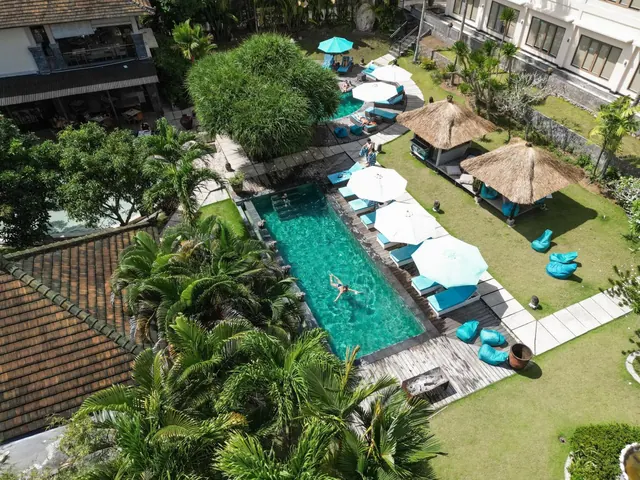 Aerial view of a long rectangular swimming pool with turquoise water, surrounded by sun loungers, white parasols, thatched pavilions and palm trees, with one swimmer in the pool.