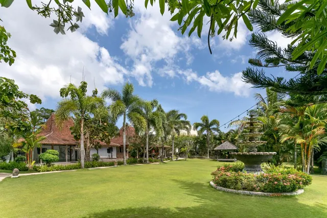 A manicured lawn with a tiered stone fountain, palm trees and a traditional tiled-roof building in the background.