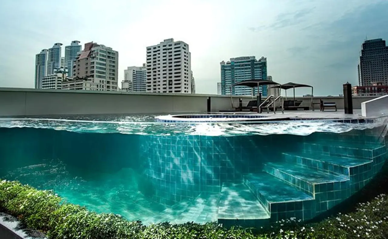 A rooftop swimming pool is shown split at the waterline, with sun loungers and umbrellas beside it and high-rise towers behind.
