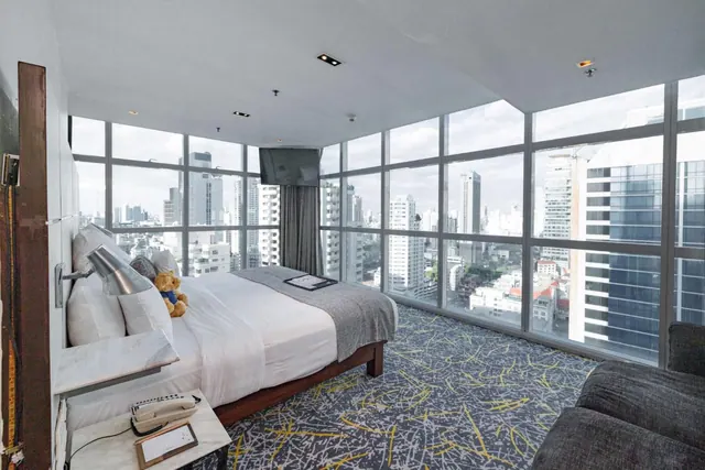 A bedroom with a made double bed and a teddy bear, with floor-to-ceiling windows looking out over high-rise buildings in the city.
