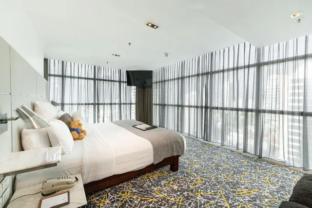 A bedroom with a large bed, a wall-mounted television and floor-to-ceiling windows with sheer curtains that look out onto tall buildings.