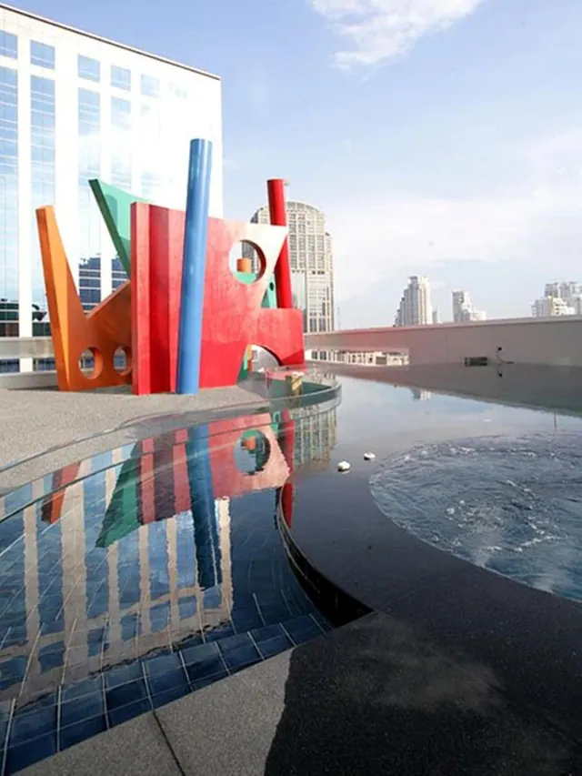 A rooftop pool with a large colorful abstract sculpture at one end and city towers in the background.