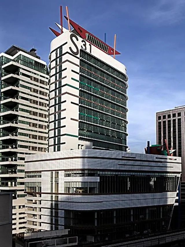A tall white and green glass tower with the S31 sign on its roof stands beside other high-rise buildings under a blue sky.