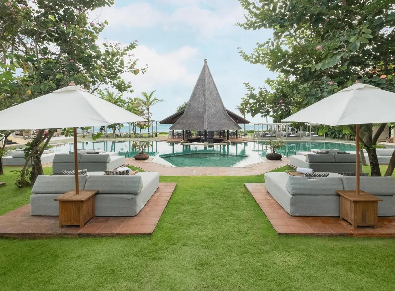A pool with a thatched pavilion at its far end, lounge sofas and white umbrellas on the grass in front, with the sea visible beyond.