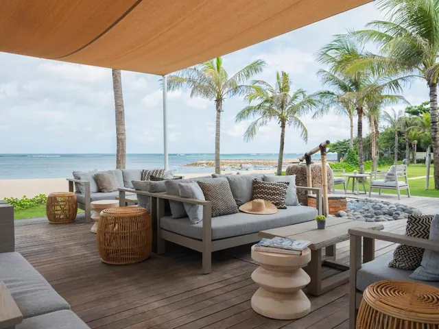 A covered wooden deck with grey outdoor sofas and rattan stools looks out over a sandy beach, the sea and palm trees.
