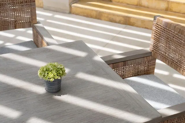 A grey outdoor table with woven rattan chairs and a small potted green plant sits in strong sunlight on a tiled terrace beside stone steps.