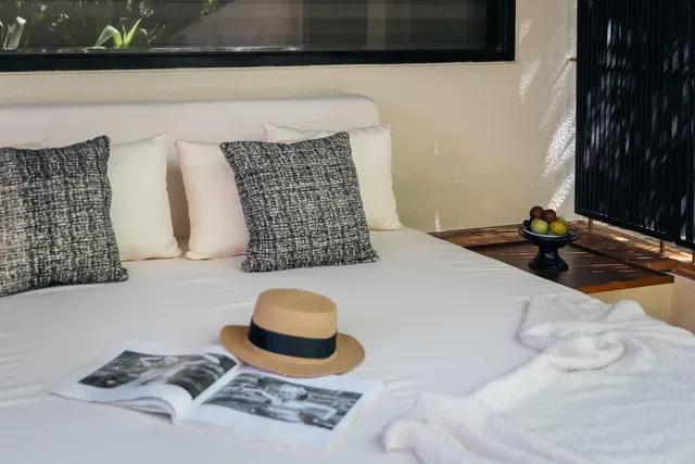 A made bed with white linens and grey patterned cushions, with a straw hat, an open magazine, a folded white towel and a fruit bowl on a wooden side table.