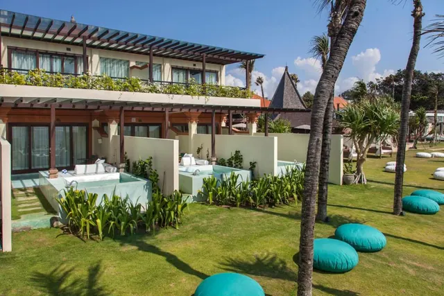 Exterior of a two-storey building with a pergola, small plunge pools in front of the ground-floor rooms, and teal cushions on a lawn with palm trees.