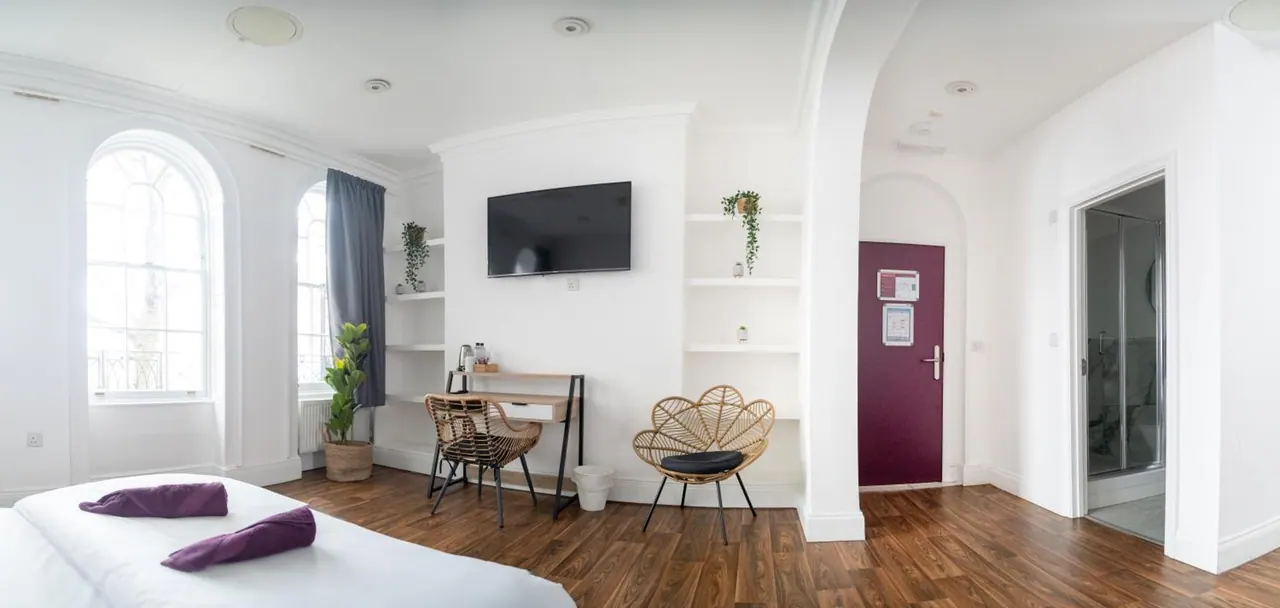 A bright white hotel room with arched windows, a bed with purple towels in the foreground, a wall-mounted TV, a wooden desk with a wicker chair, and a glass-walled shower to the right.