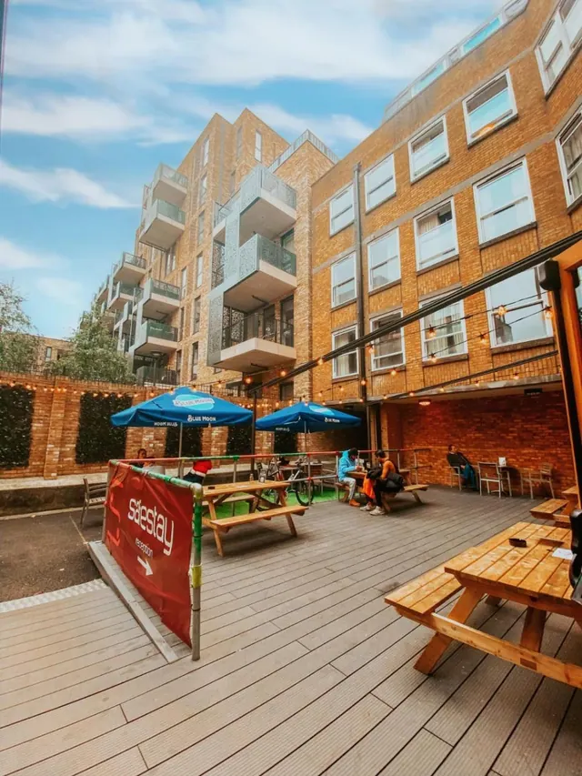 An outdoor courtyard terrace with wooden picnic tables under blue Blue Moon umbrellas on a timber deck, with brick apartment buildings behind and a red Safestay banner at the left.