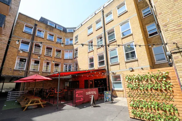 A paved courtyard with wooden picnic tables under red umbrellas and a red awning, surrounded by yellow brick buildings with string lights strung overhead.