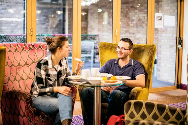 Two people sit in patterned armchairs at a small glass table with plates, cups and orange juice, with a brick courtyard visible through glass doors behind them.