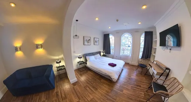 A wide-angle view of a bedroom with a double bed, a blue sofa, a wall-mounted television, a work desk and tall arched windows.