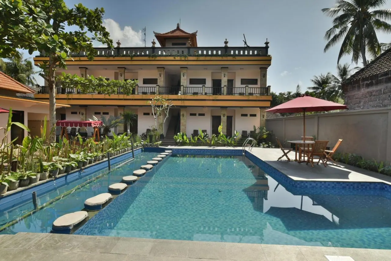 A swimming pool with stepping stones sits in front of a yellow and white multi-storey building with balconies, with a red parasol and table set to the right.