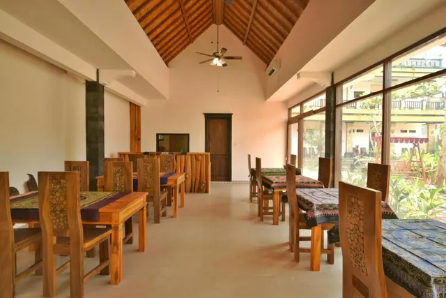 A dining room with wooden tables and chairs, batik-covered tables, a timber vaulted ceiling and large windows looking onto a garden.