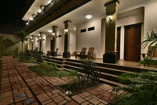 A night view of a columned veranda with wooden doors, two wooden chairs on a raised terrace, and a brick path lined with plants.