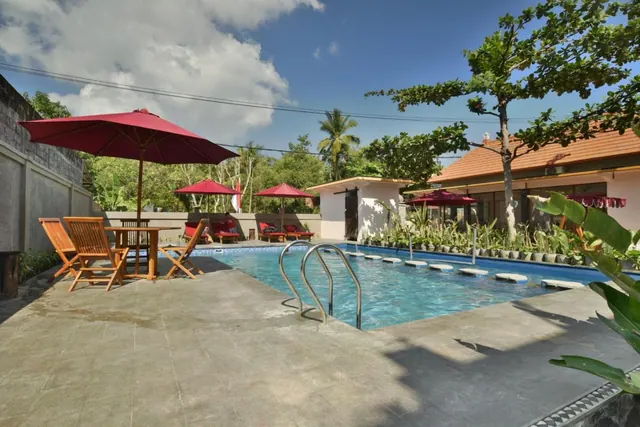 A small swimming pool with metal handrails and stepping stones sits beside a tiled terrace with red-umbrella loungers and a wooden table set.