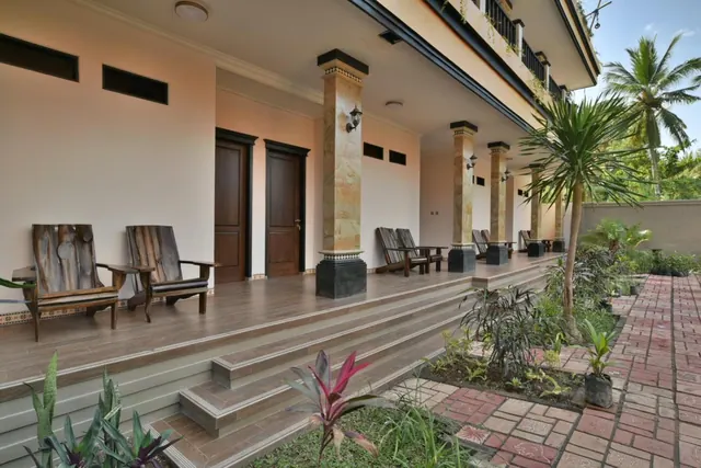 A covered veranda with carved wooden armchairs, stone columns, doors to rooms, and a stepped walkway leading to a brick path lined with plants.